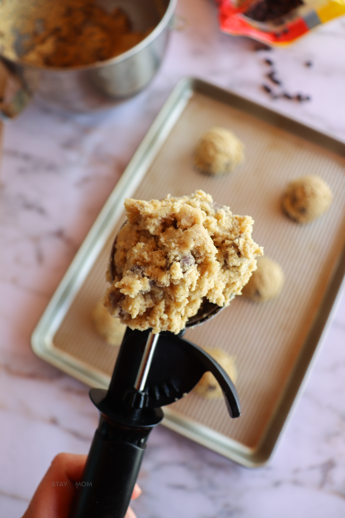 chocolate chip cookie dough on a scooper with the sheet pan behind it with more cookie dough balls on it.