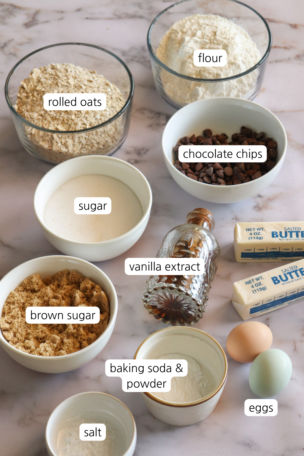 All the ingredients to make oatmeal chocolate chip cookies in bowls. Oats, flour, chocolate chips, sugar, brown sugar, vanilla extract, butter, eggs. salt, baking powder and baking soda.