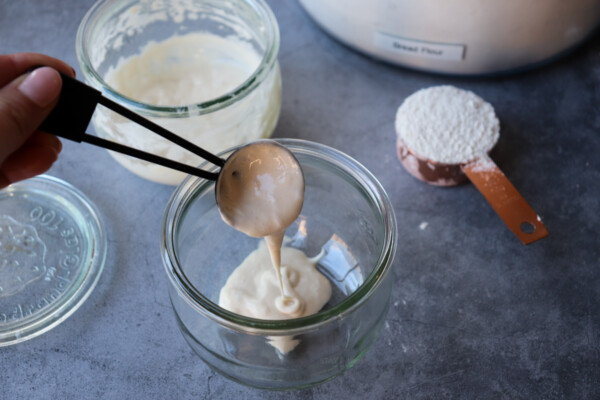 pouring sourdough starter into a clean glass jar with flour in a measuring spoon