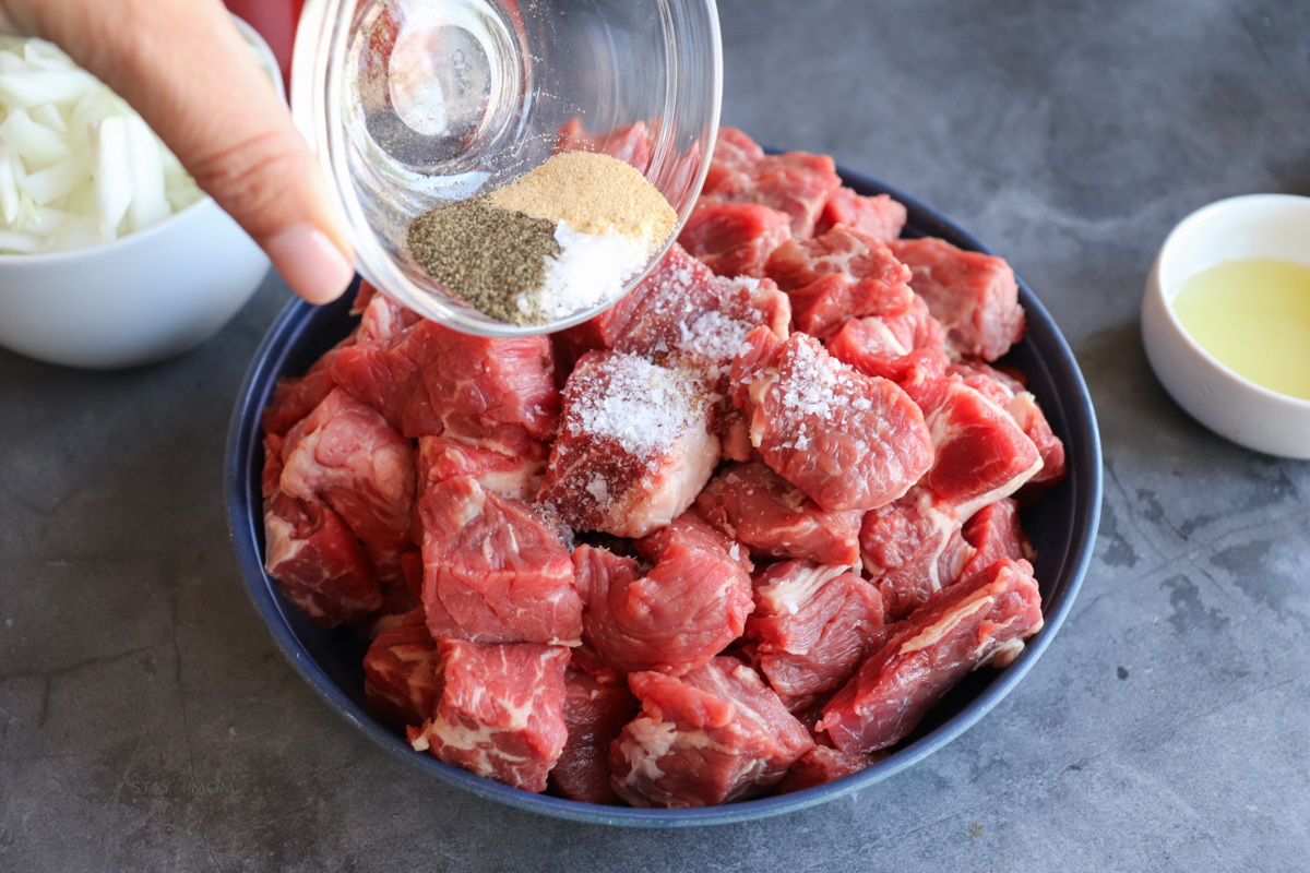 Texas chili showing bowl of diced chuck roast being sprinkled with seasonings from the recipe.