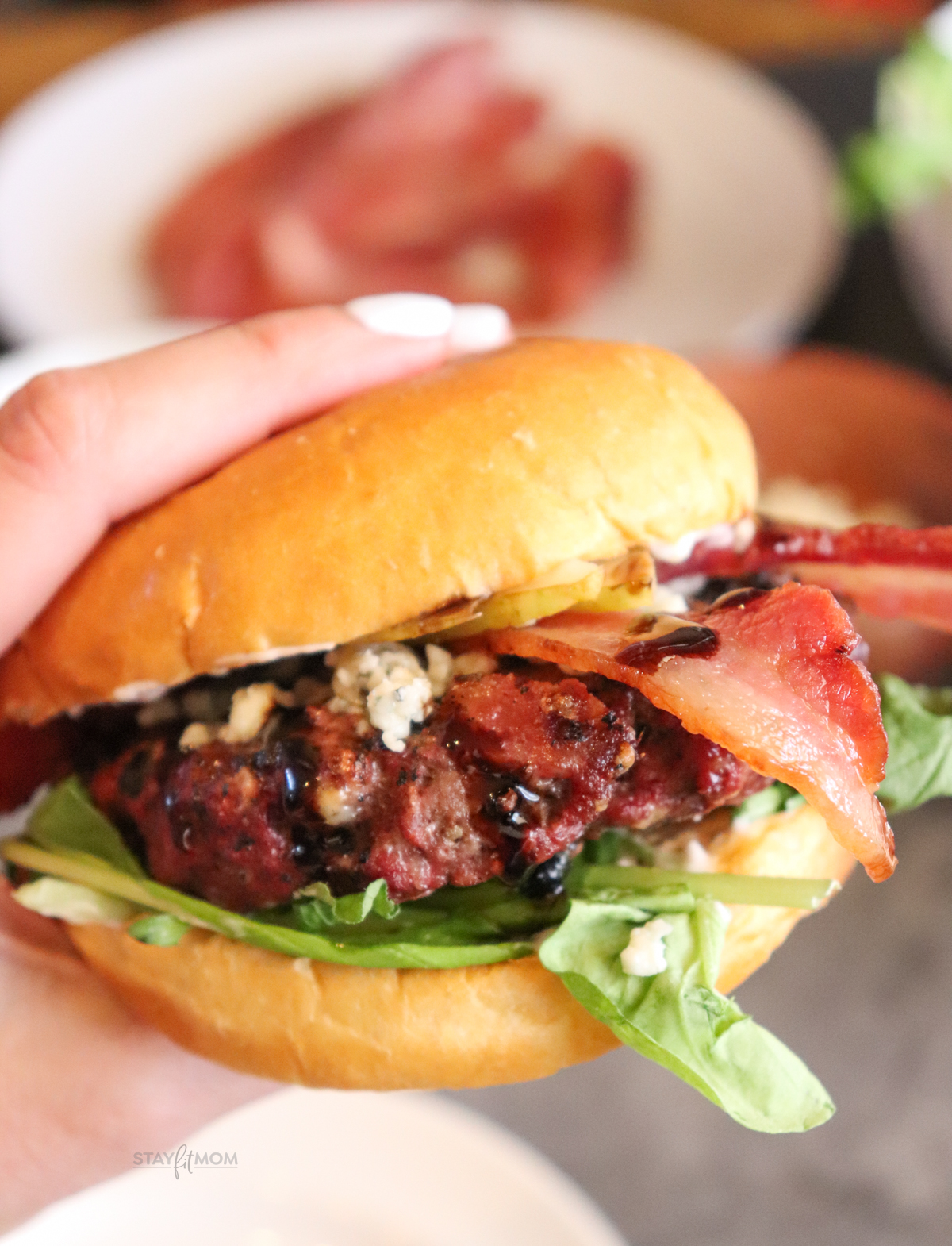 Blue Cheese & Bacon Burgers showing up close image of completed burger with visible grilled burger, arugula, cooked bacon, and blue cheese crumbles.