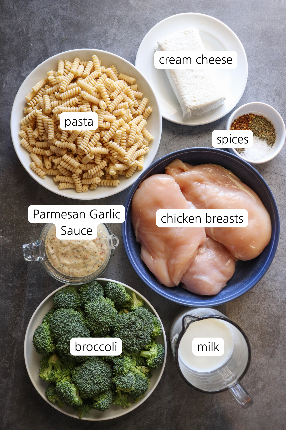 Overhead view of recipe ingredients including raw chicken breast, Buffalo Wild Wings Parmesan Garlic sauce, milk, cream cheese, pasta, and broccoli florets.
