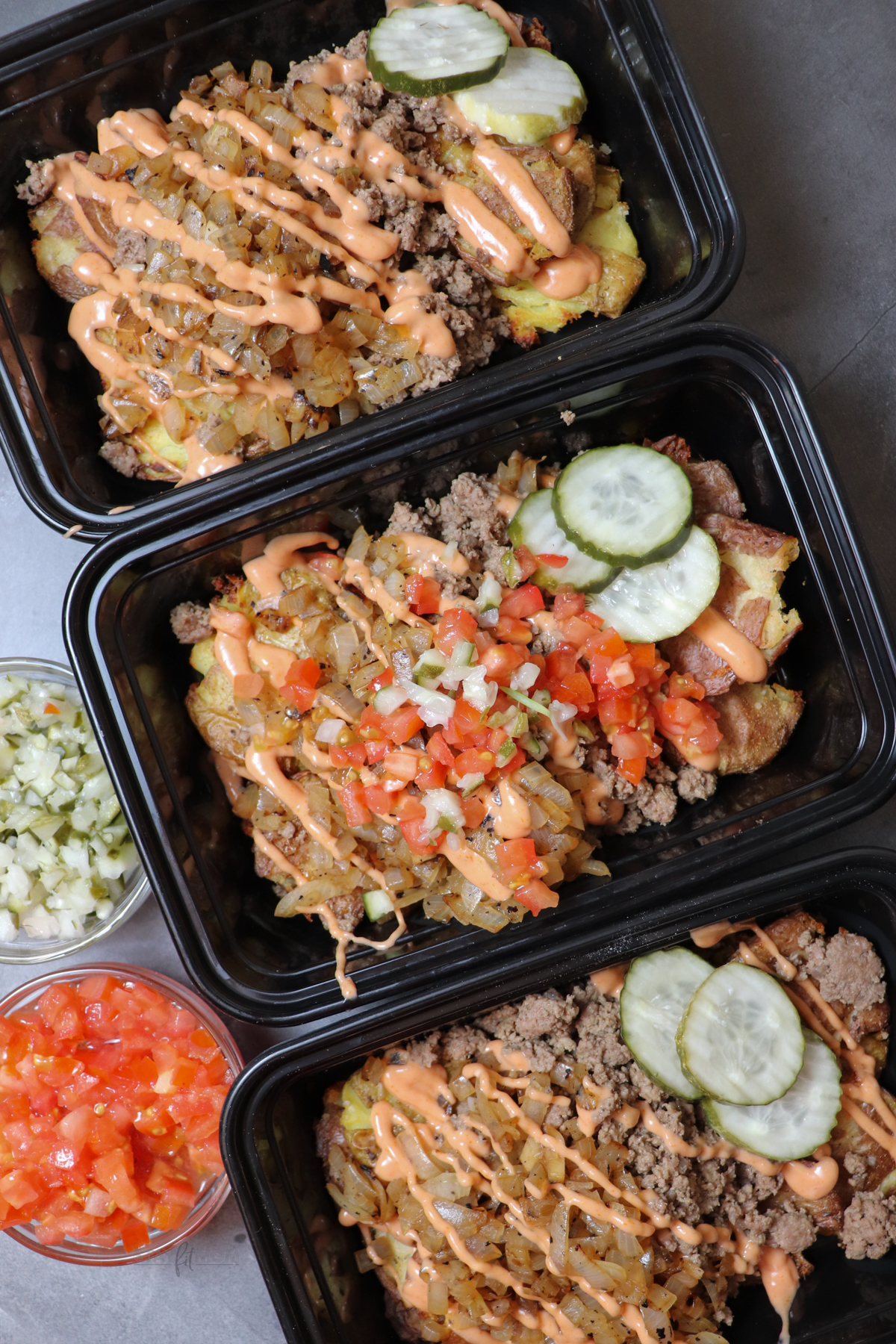 Animal style burger bowls showing meal prep containers loaded with smashed potatoes, ground beef, caramelized onions, topped with pickles, diced onions, tomatoes, and sauce.