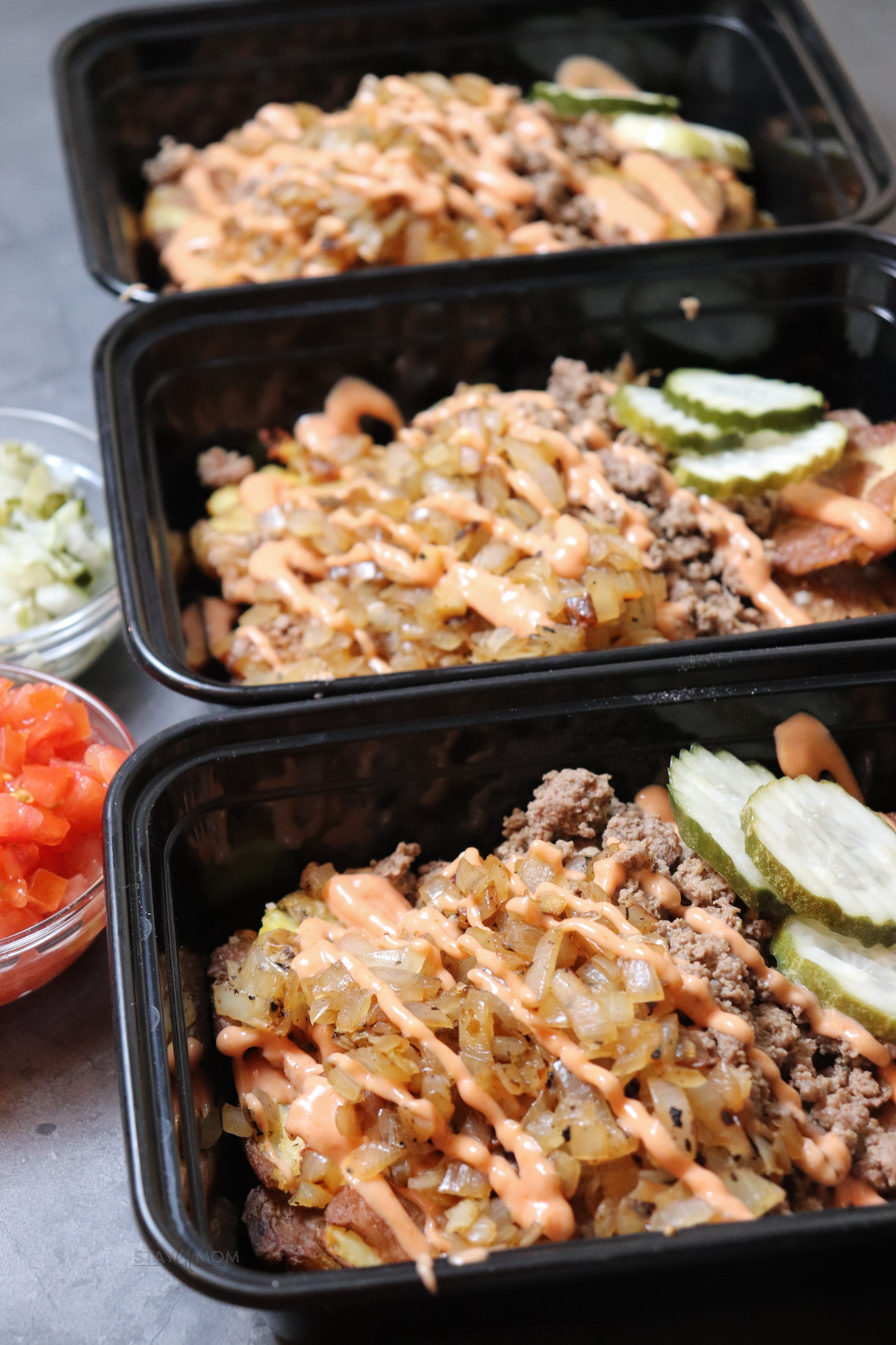 3 meal prep containers lined up and filled with crispy potatoes, ground beef, caramelized onions, and topped with pickles and sauce.