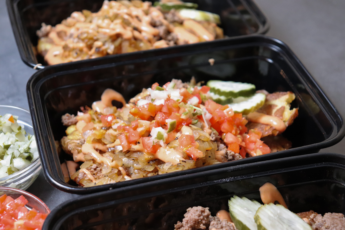 Animal style burger bowls showing meal prep containers loaded with smashed potatoes, ground beef, caramelized onions, topped with pickles, diced onions, tomatoes, and sauce.
