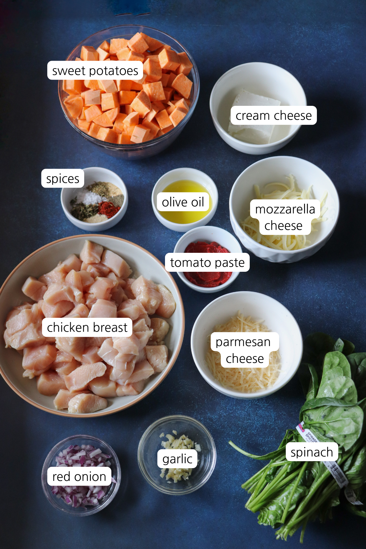 Small bowls with labeled ingredients including: sweet potatoes, cream cheese, spices, olive oil, shredded mozzarella cheese, tomato paste, parmesan cheese, chicken breast, spinach garlic, and red onion.