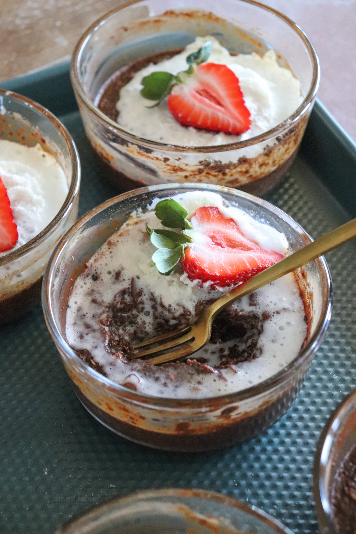 Chocolate baked oats showing completed recipe with glass meal prep bowls filled with baked oats, topped with chocolate, whipped cream, and a strawberry slice. with a bite missing and a fork in the dish.