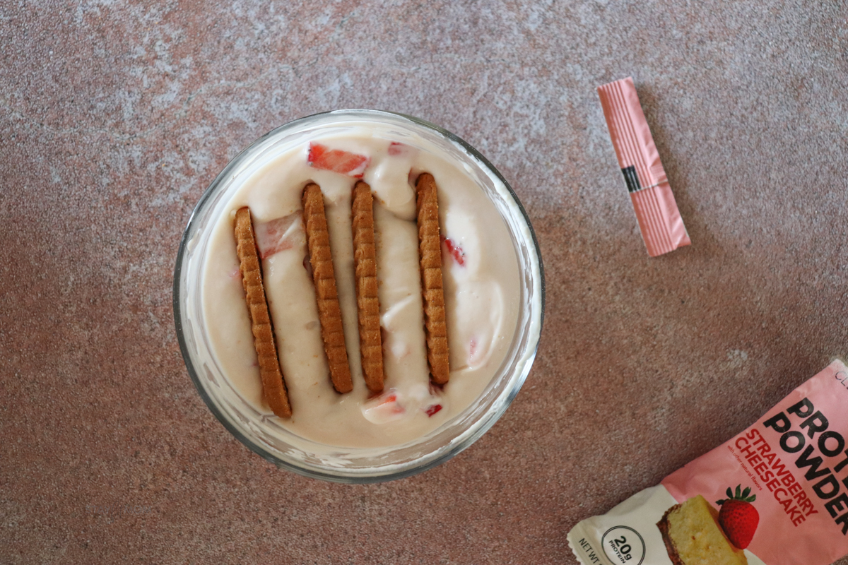 Small glass bowl filled with a mixture of Greek yogurt, protein powder, and strawberries with 4 biscoff cookies lined up and pushed into the mixture.
