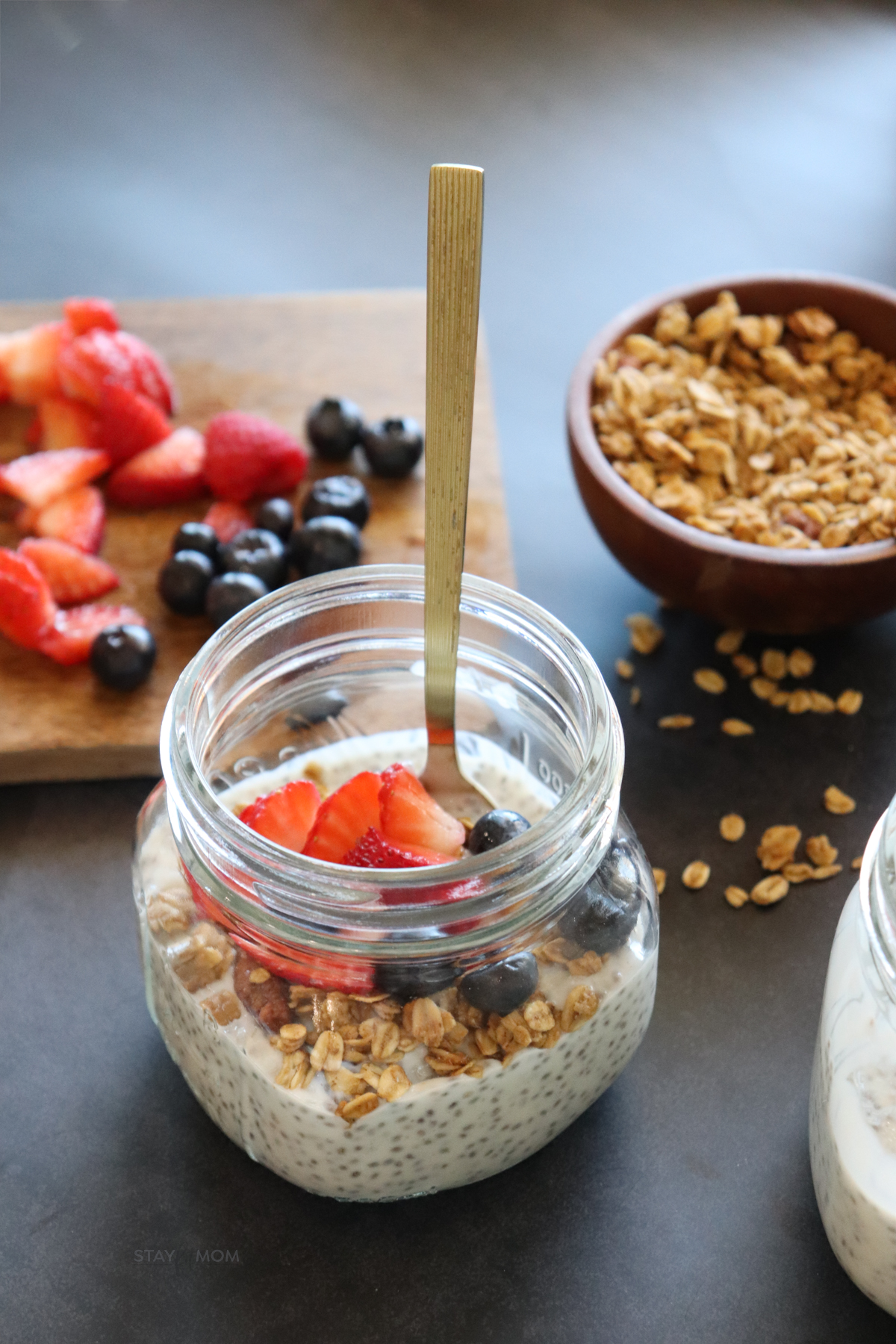 Vanilla Chia Pudding showing completed recipe in a small jar topped with fresh berries and granola with a spoon in the jar and fresh berries and granola in the background.