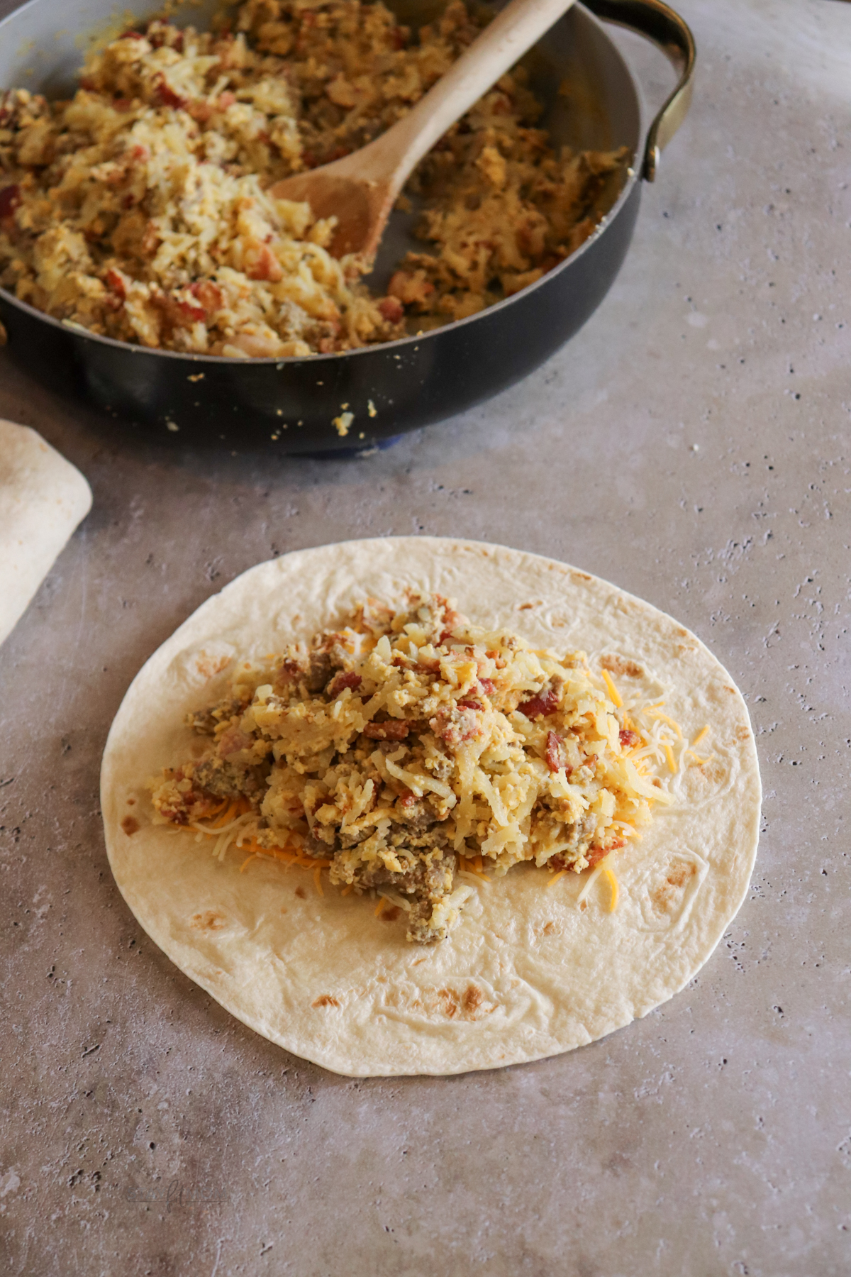 Macho Breakfast Burritos showing a tortilla with the filling ready to be rolled with a pan of filling in the background.