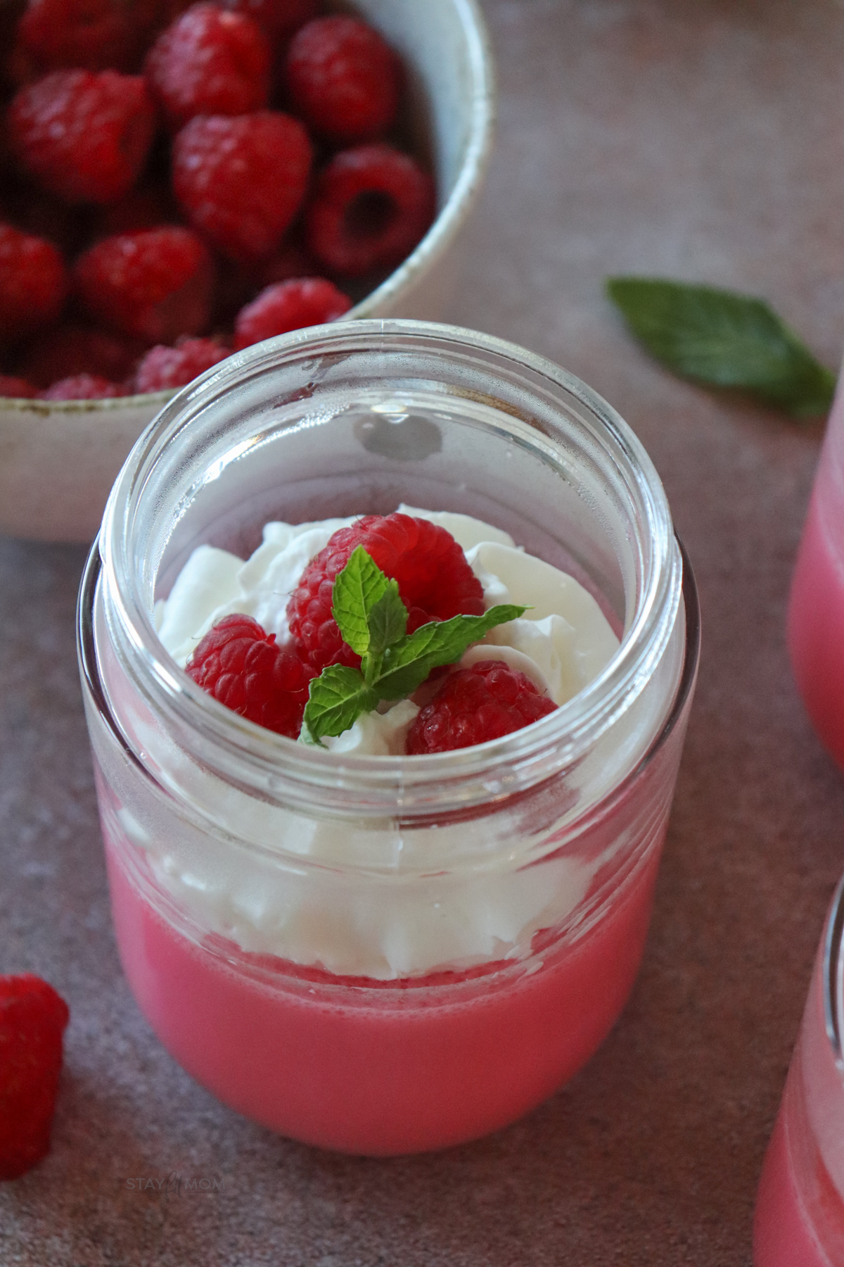 Yogurt Jello cups showing a small glass jar filled with the mixture, topped with whipped cream, fresh raspberries, and a sprig of mint.