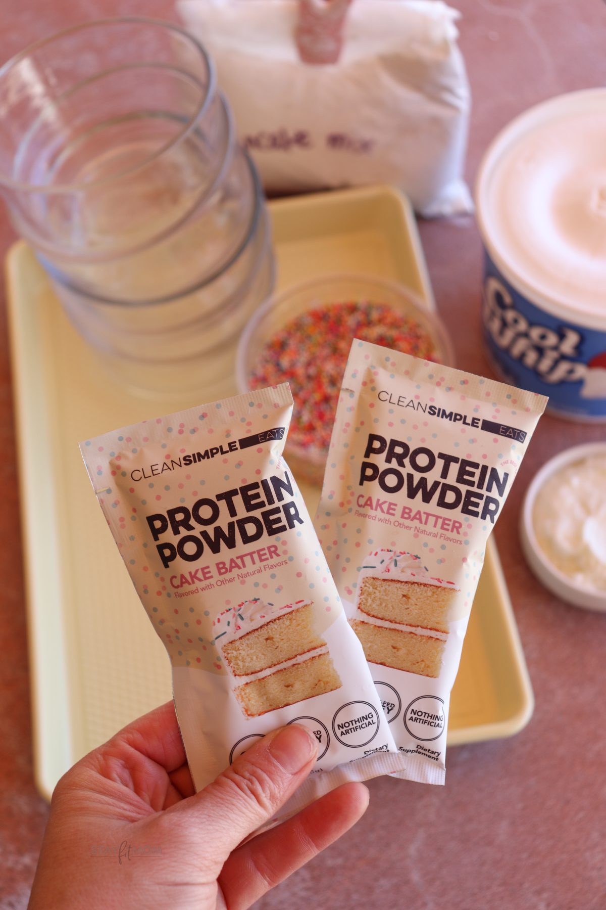 Two packages of Clean Simple Eats cake batter protein being held above a tray with glass meal prep bowls, sprinkles, and Cool Whip.