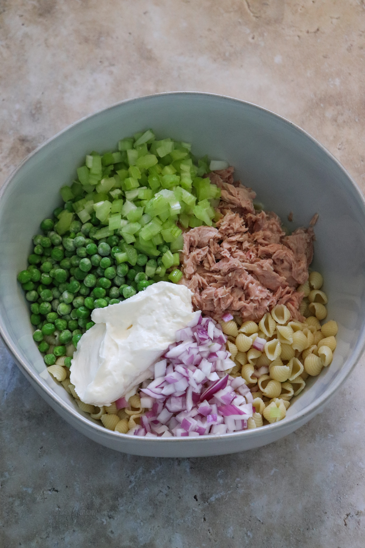 Tuna pasta salad showing a mixing bowl with cooked pasta, tuna chopped purple onion, pease, chopped celery, and Greek yogurt ready to be stirred together.
