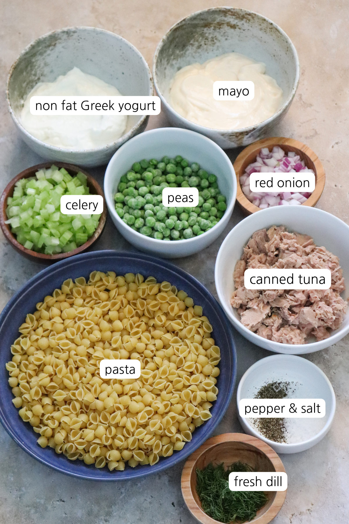 Tuna Pasta salad showing ingredients labeled in small bowls: shell pasta, fresh dill, salt & pepper, canned tuna, red onion, pease, celery, may, and non fat Greek yogurt.