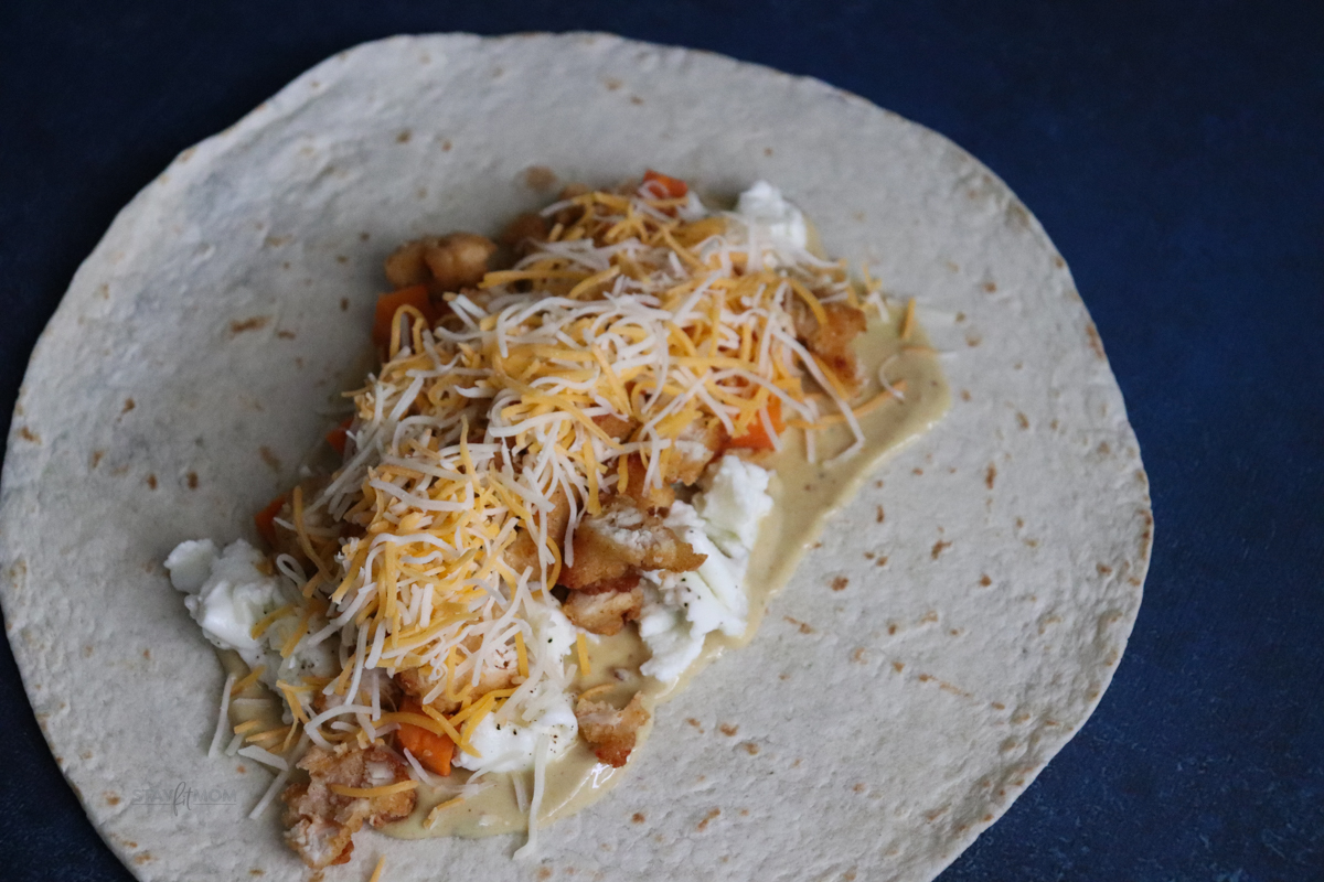 Tortilla filled with sauce, sweet potatoes, scrambled egg whites, crispy chicken, and cheese ready to be rolled into a burrito,