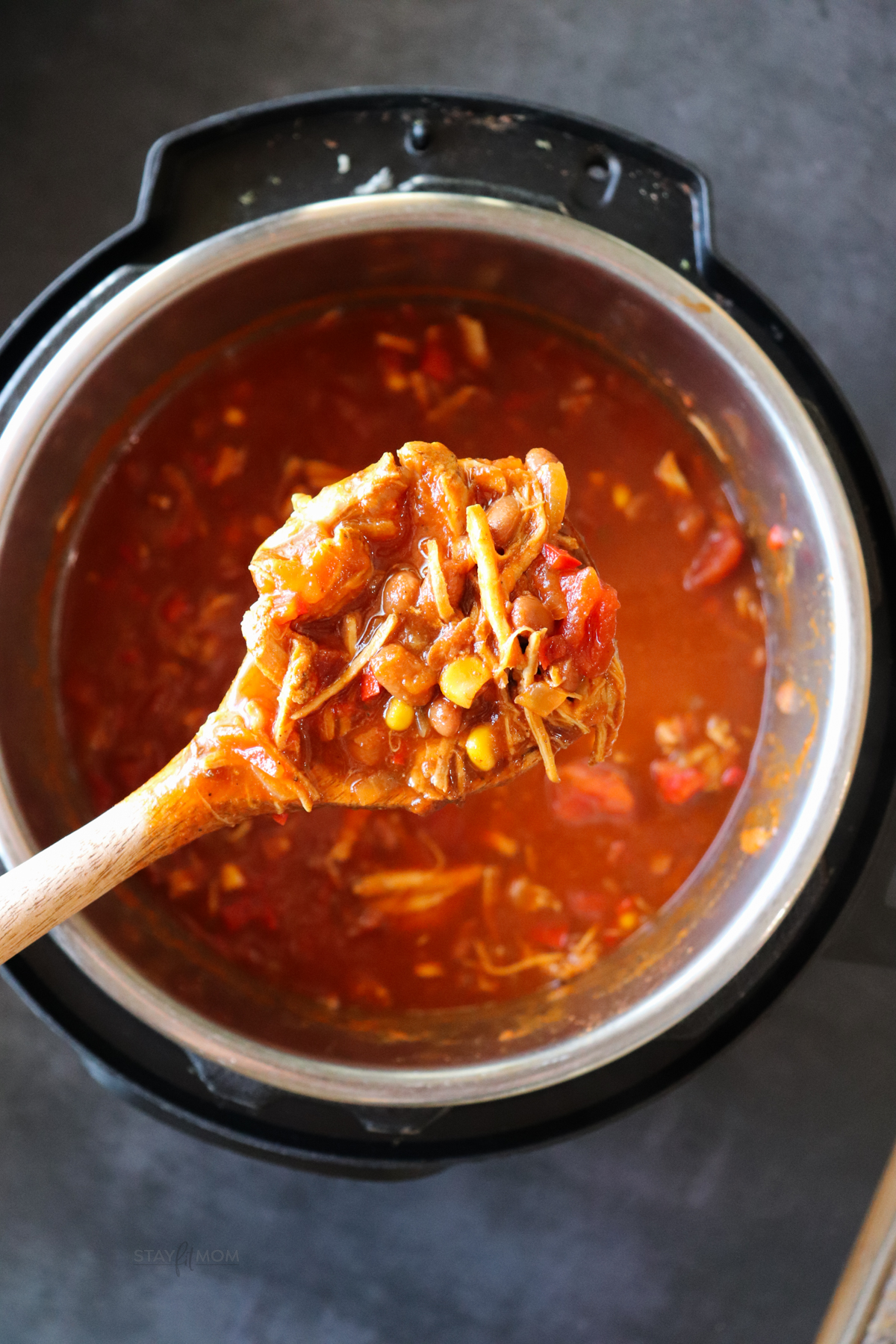 Instant Pot filled with Pulled Pork Chili with a wooden spoon being held above with a scoop of the chili to show shredded pork, tomatoes, corn, sauce, etc.