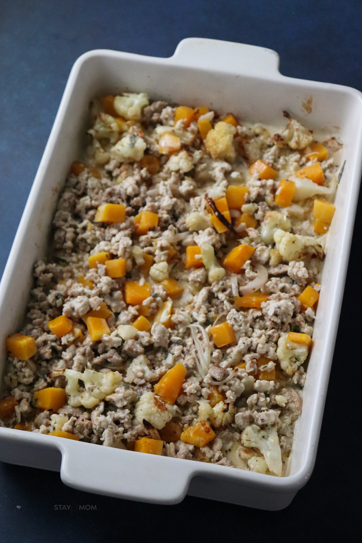 A white 9x13 pan filled with butternut squash, cauliflower florets, onions, sausage, and spices ready to be topped with cheese.