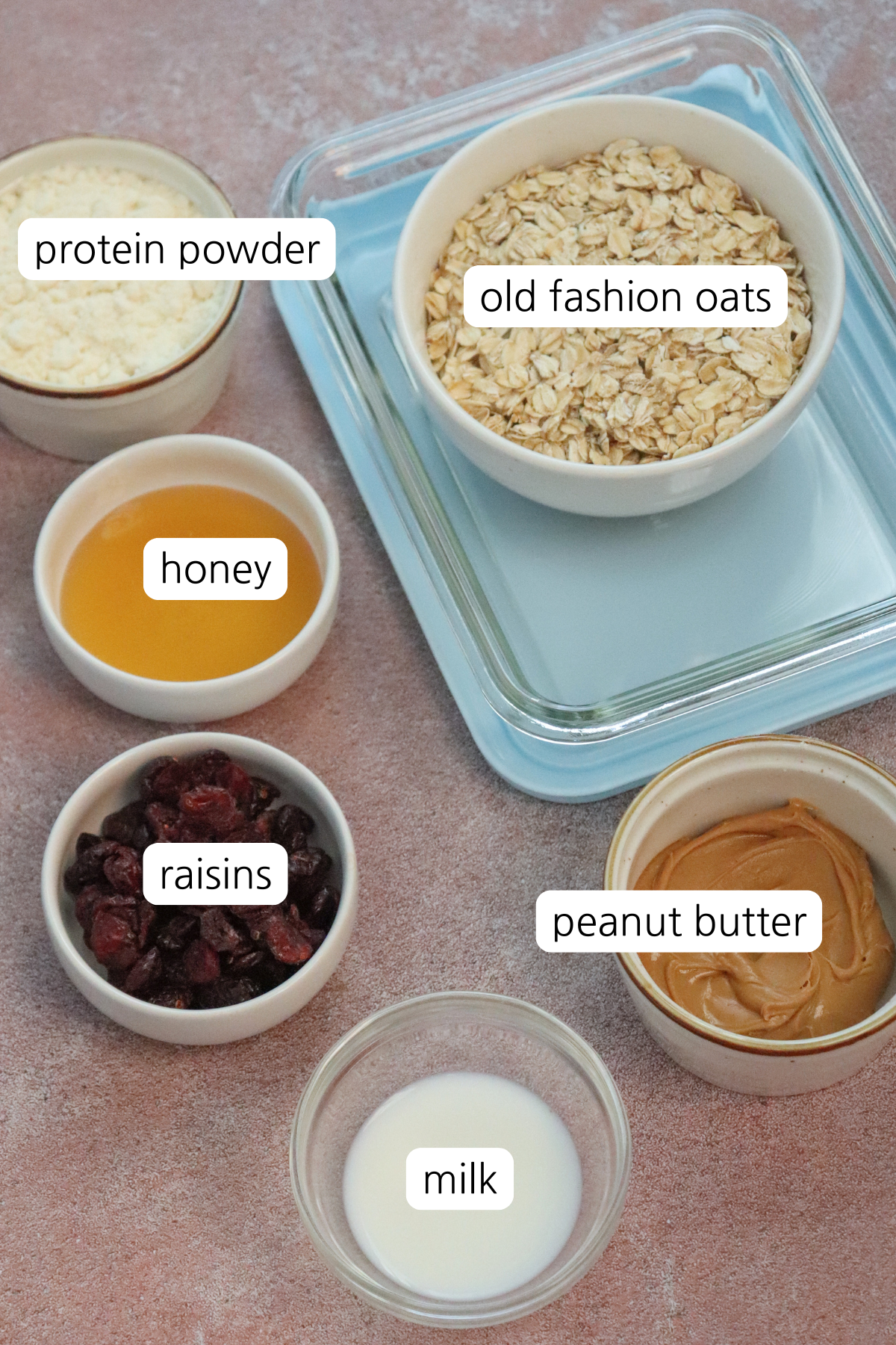 PB&J Protein bites showing ingredients labeled in small containers: protein powder, old fashioned oats, honey, raisins, peanut butter, and milk.