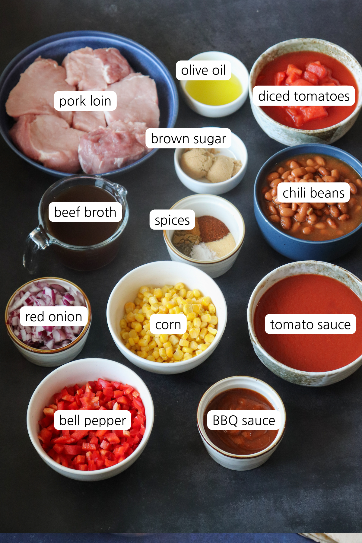 Pulled Pork Chili showing small bowls labeled with ingredients: pork loin, olive oil, died tomatoes, brown sugar, chili beans, spices, beef broth, red onion, corn, tomato sauce, bell pepper, and BBQ sauce.