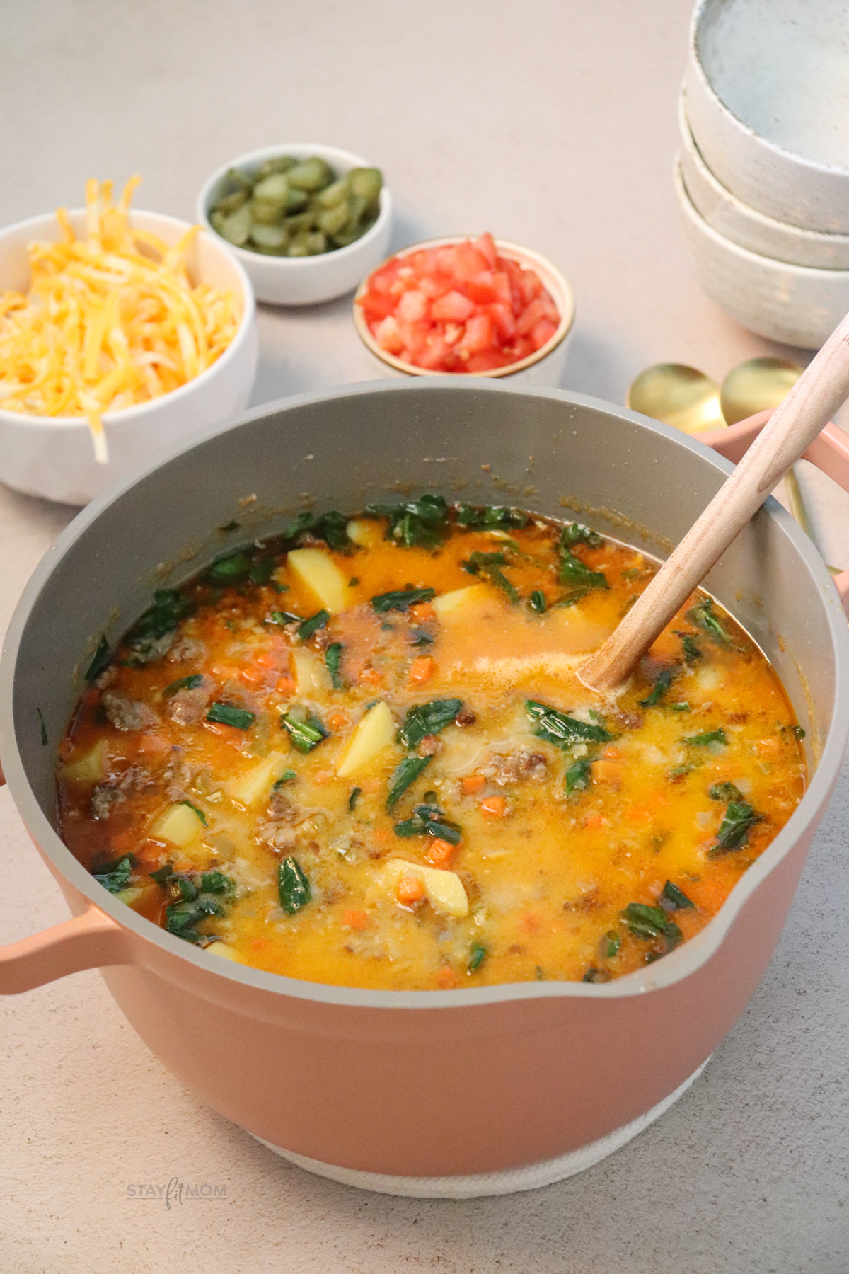 A pot of Cheeseburger soup with cheese, tomatoes, and pickles in the background.