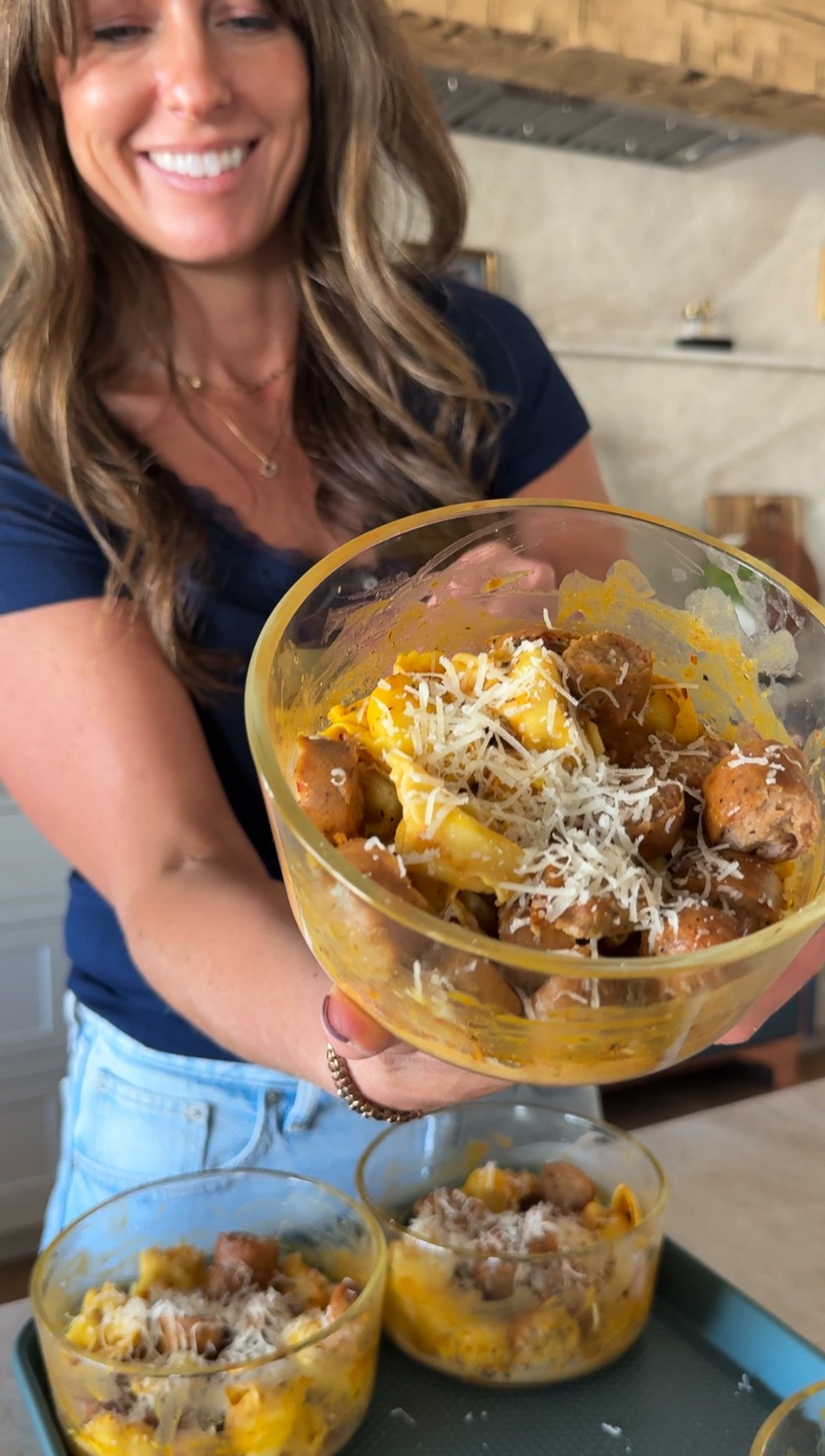 Pumpkin Tortellini Bowls with Krista showing a finished meal prep recipe to the camera with visible tortellini, chicken sausage, sauce, and topped with Parmesan cheee.