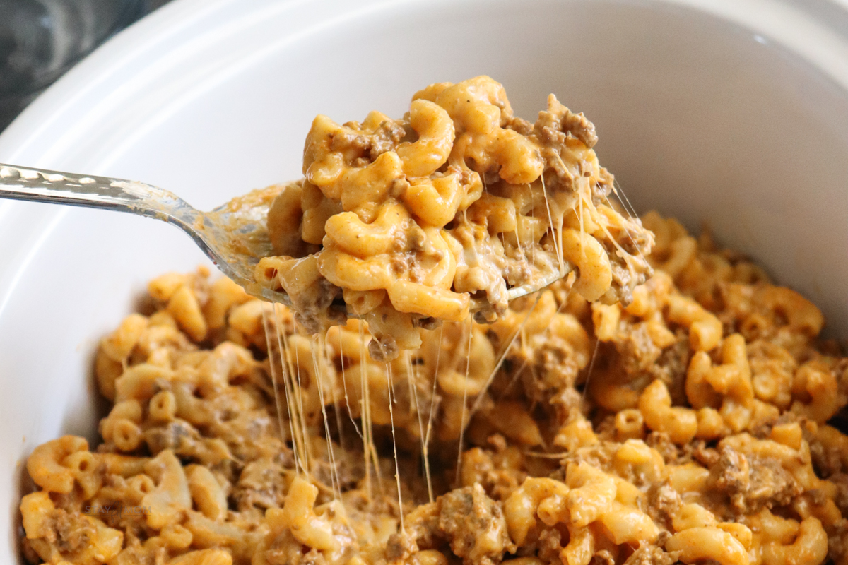 A close up image of Hamburger Mac showing finished recipe in a white crock with a spoon scooping some up and a cheese pull.