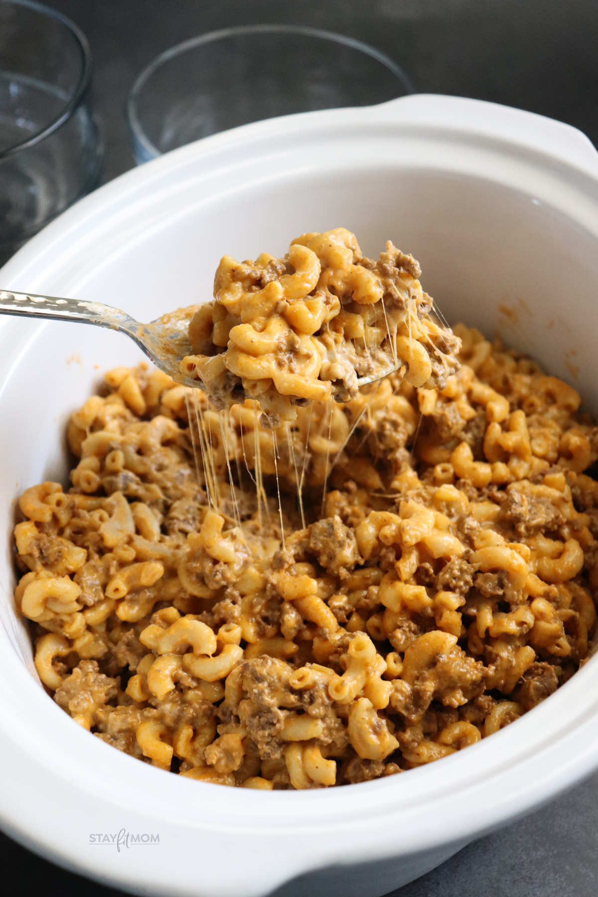 Hamburger Mac showing finished recipe in a white crock with a spoon scooping some up and a cheese pull.