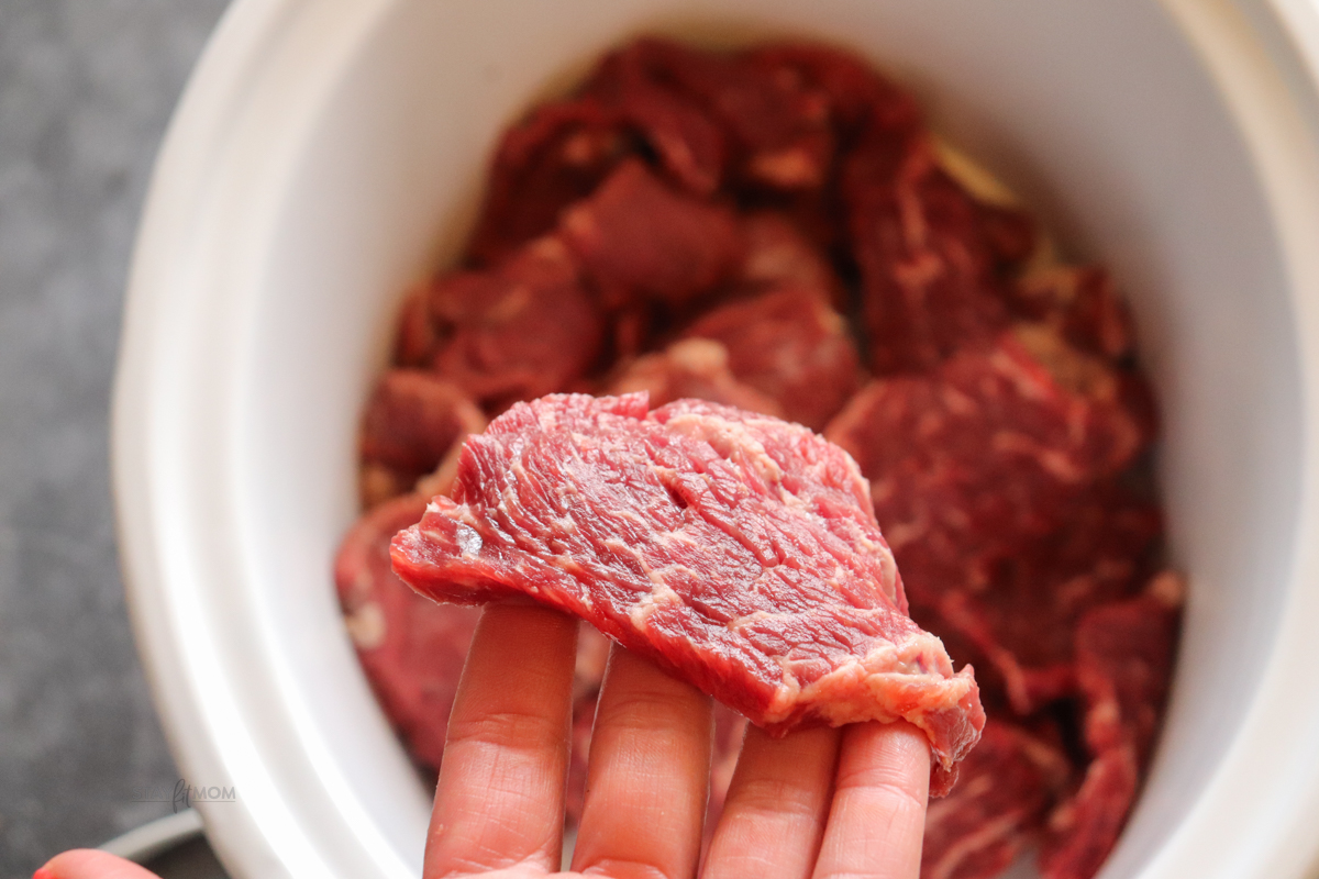 Slow cooker filled of sliced chuck roast with a piece being held above to show size.