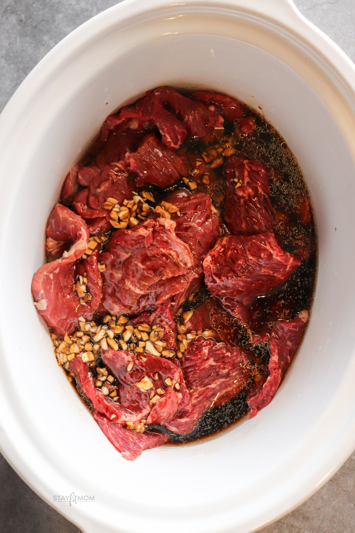 Slow cooker filled with sliced chuck roast in sauce and topped with garlic ready to be cooked.