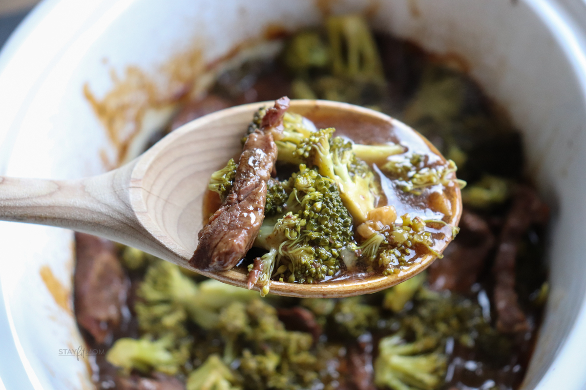 Slow cooker showing finished recipe with a wooden spoon held above showing cooked chuck roast, broccoli, and sauce.
