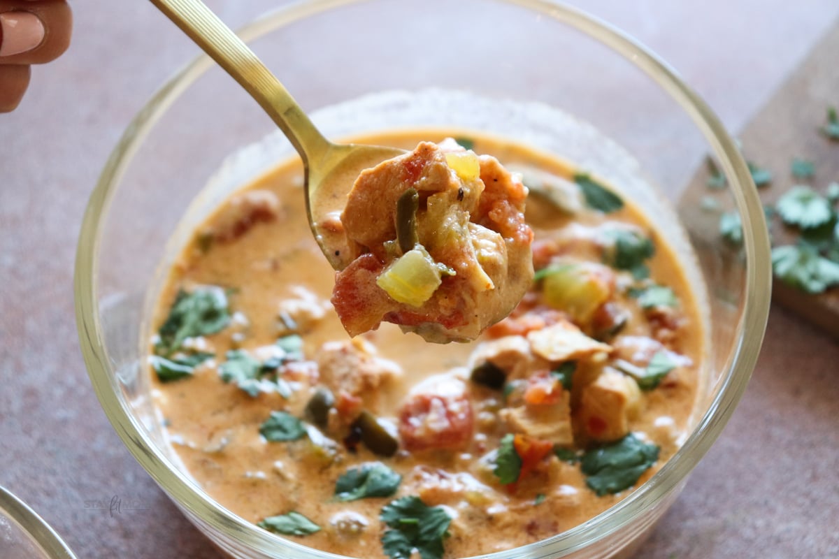A glass meal prep bowl with a serving of soup with a gold spoon held above to show texture.